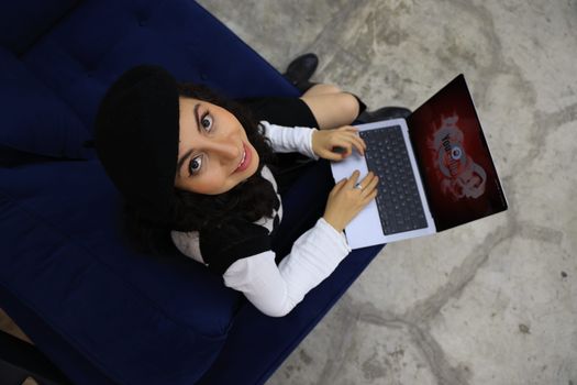 Young woman in a beret smiling while using a laptop on a couch, viewed from above.