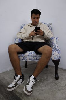 A young man sits comfortably on an armchair, focused on his smartphone, wearing a hoodie and sneakers.