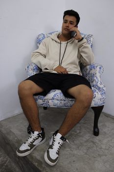 Adult man in a beige hoodie sits on a blue-patterned armchair indoors while talking on his phone.