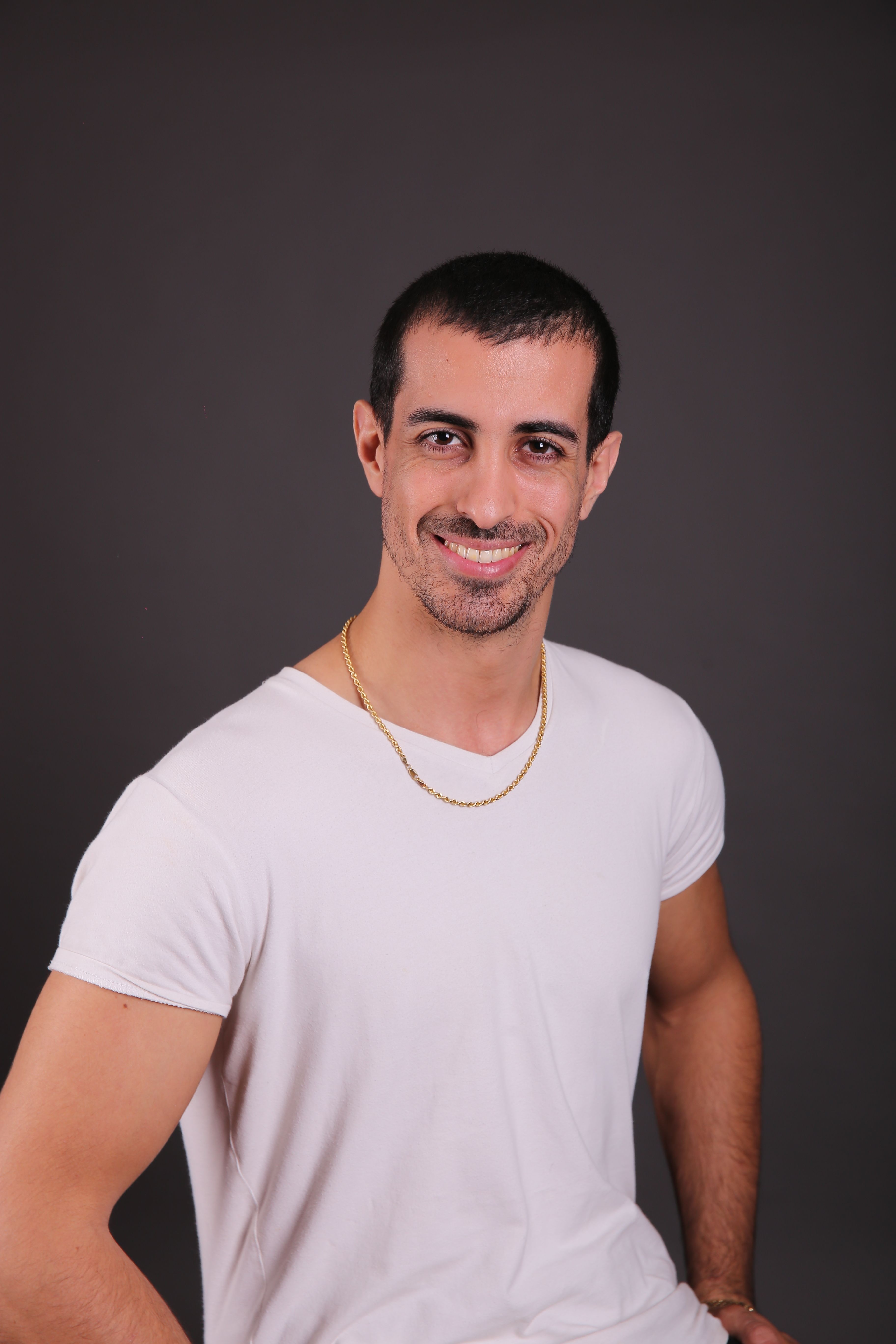 A confident young man in a casual white T-shirt smiles warmly in a studio setting.