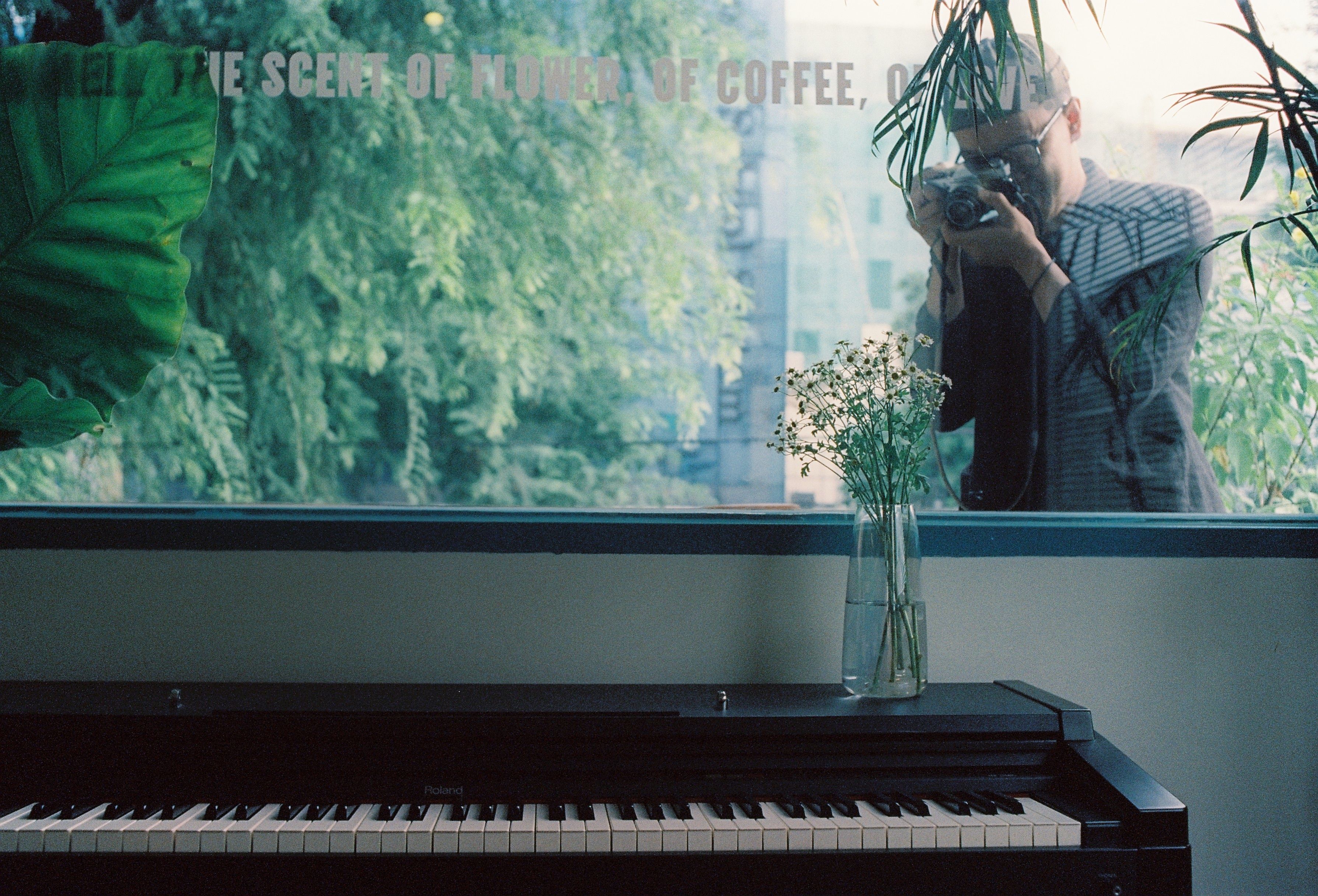A man captures a photo through a window, with a piano and vase in the foreground.