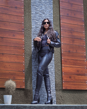 Confident woman posing in leather jacket and boots against modern urban wall.