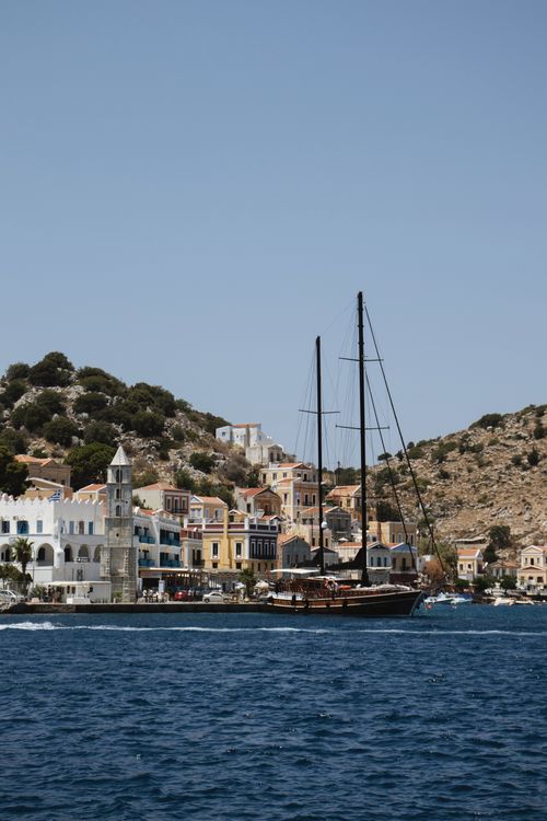 Free Explore the serene beauty of Symi's harbor with its colorful houses and classic sailboat in Greece. Stock Photo