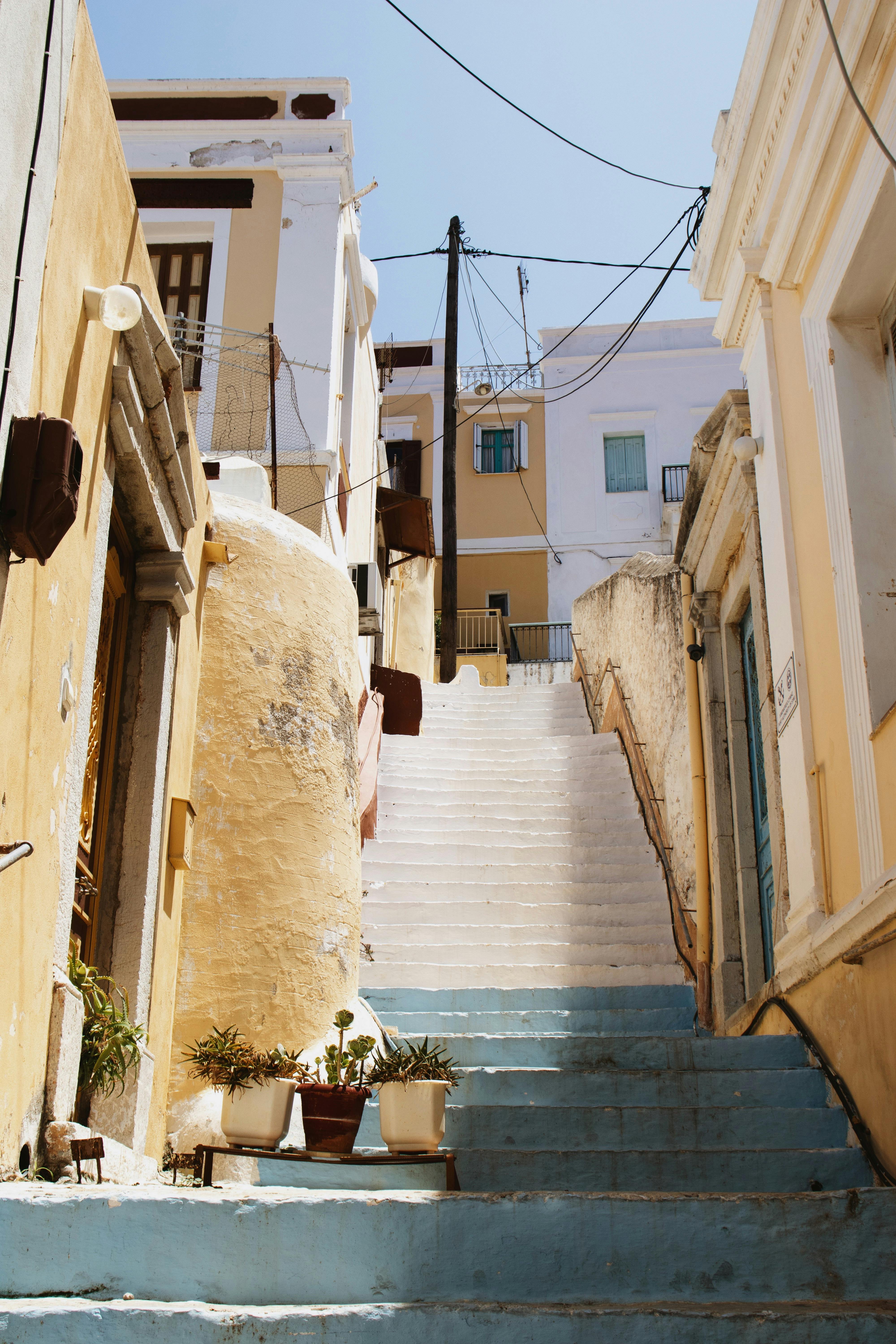 Explore a sunlit alley in a Greek town, featuring traditional staircases and vibrant architecture.