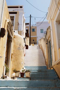 Explore a sunlit alley in a Greek town, featuring traditional staircases and vibrant architecture.