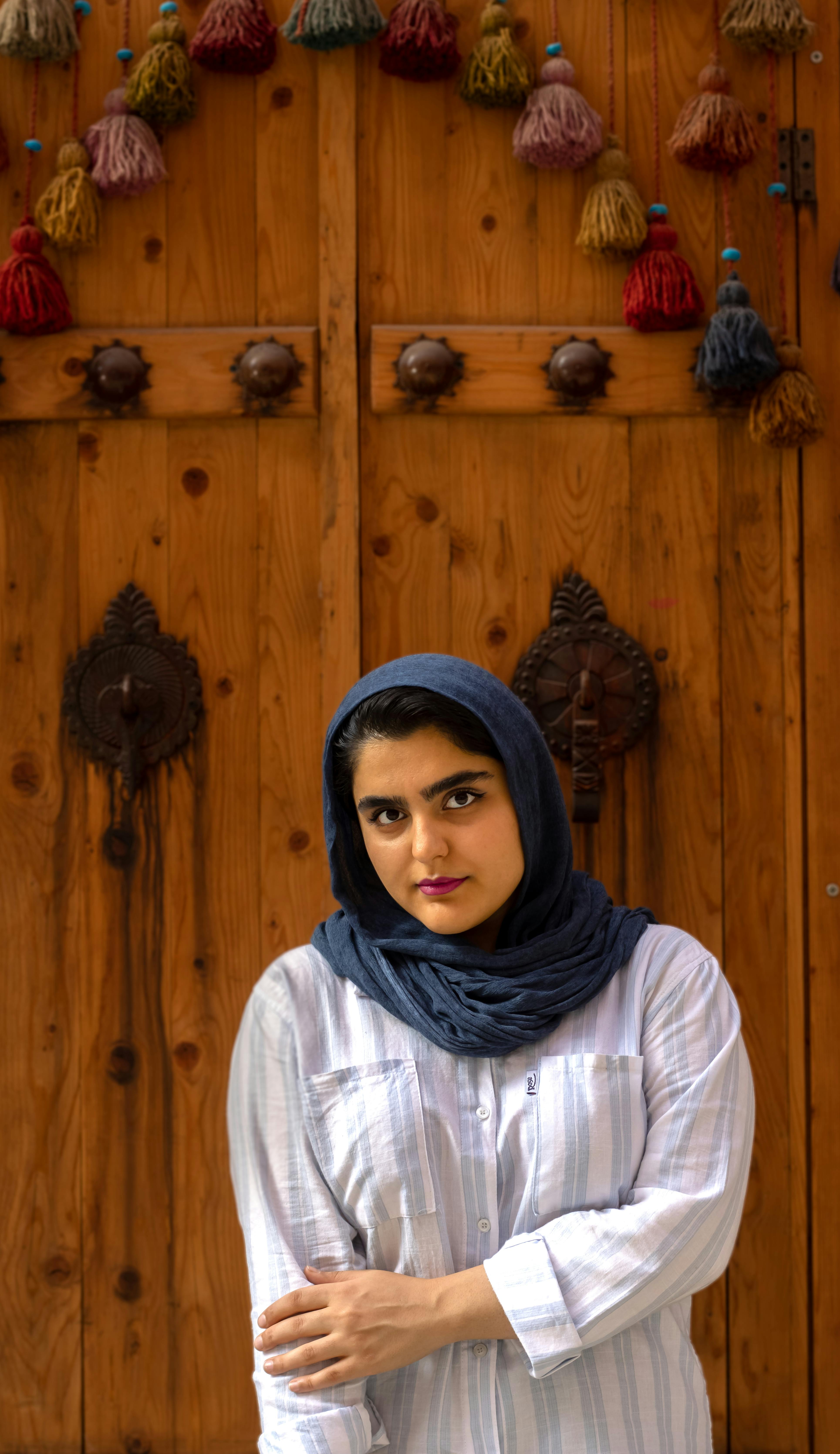 Portrait of a young woman in a hijab standing by a traditional wooden door with colorful tassels.