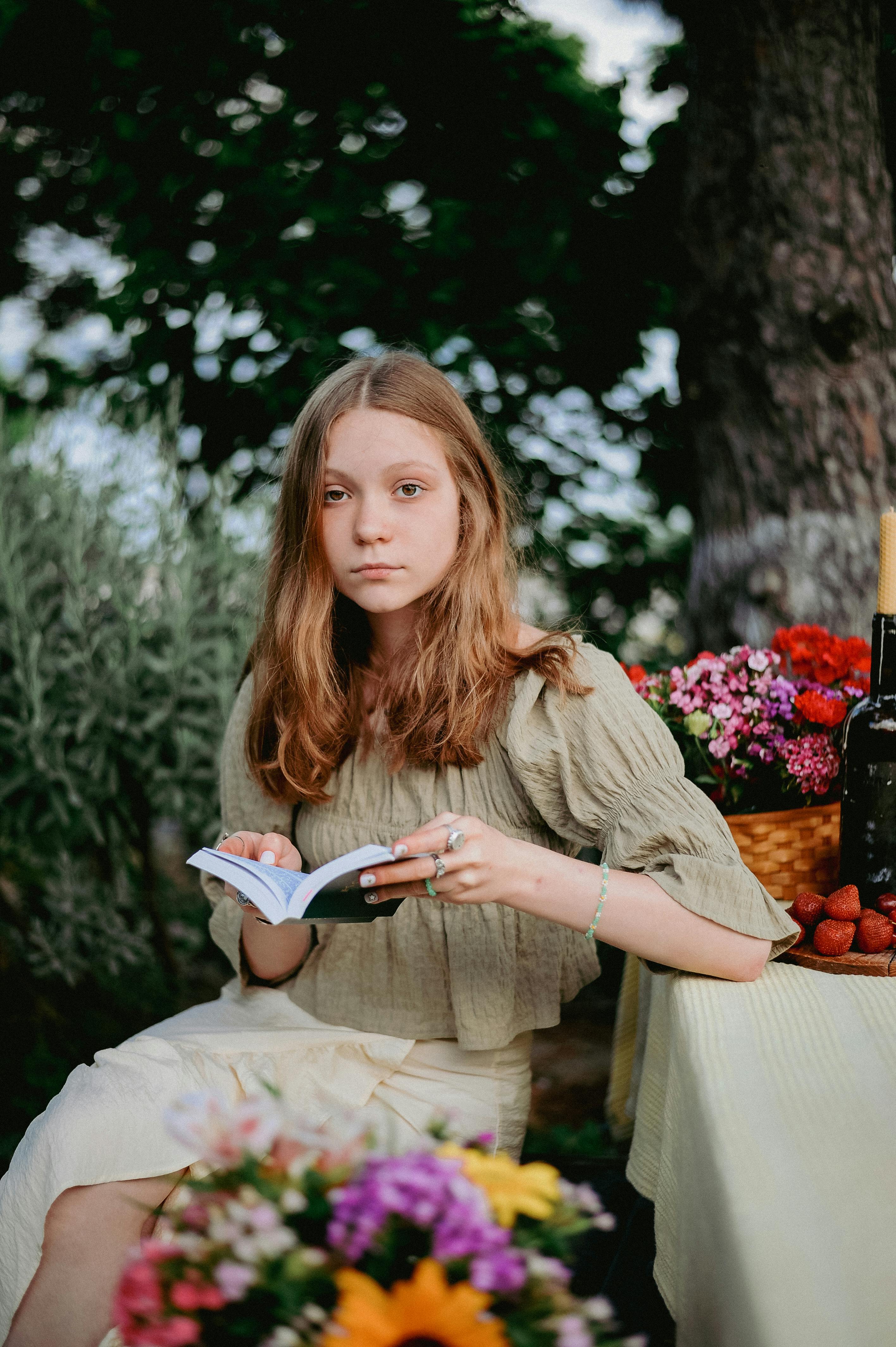 A teenage girl with long hair enjoying leisure time reading at a picnic table surrounded by summer blooms.