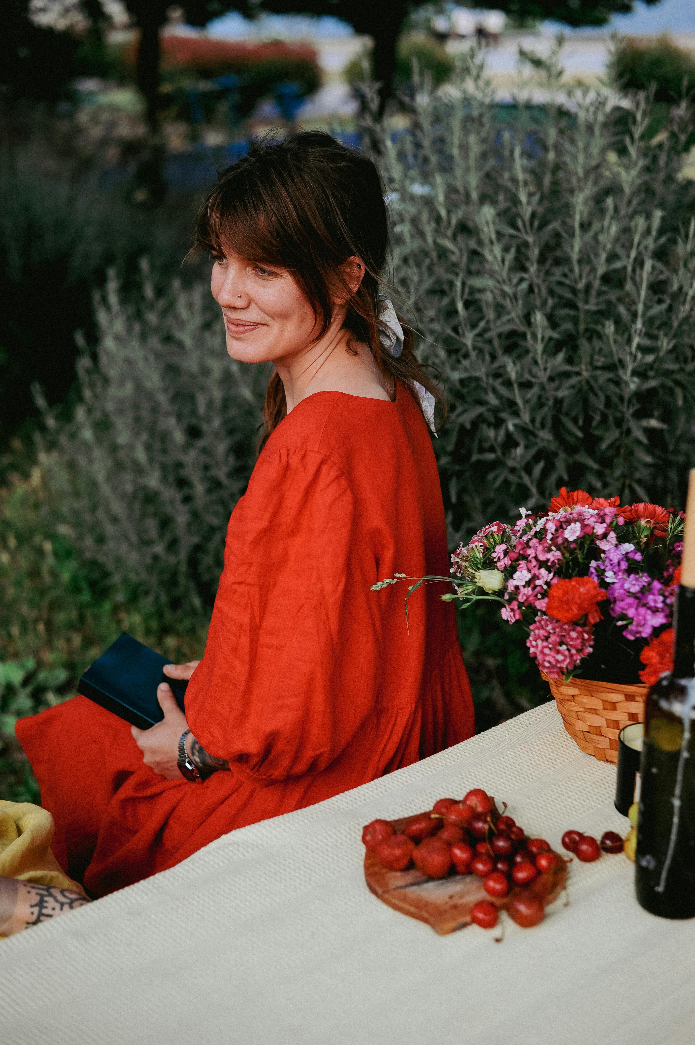 Young woman in red dress sitting by a table with flowers and cherries outdoors.