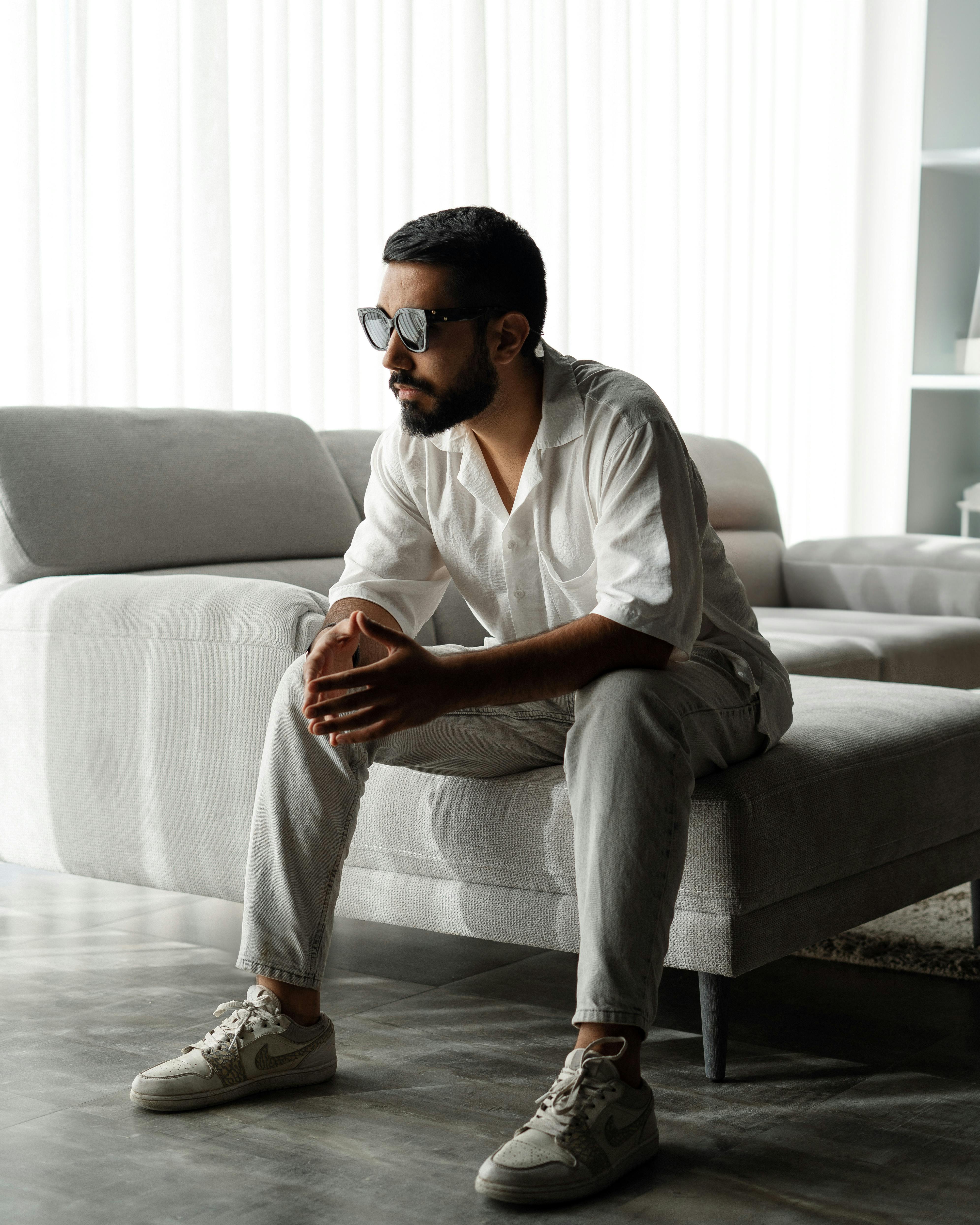 A well-dressed man in sunglasses sits on a modern sofa in a minimalist, sunlit room.