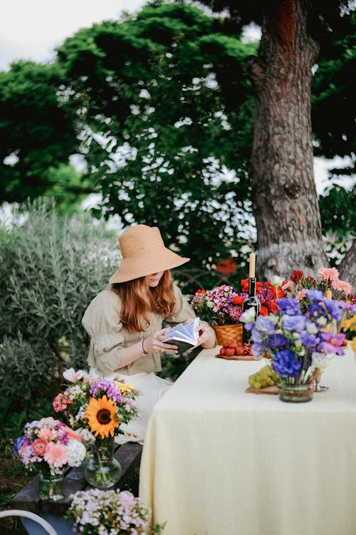 Young Woman Sitting at the Table in the Garden Decorated with Flowers and Reading a Book 