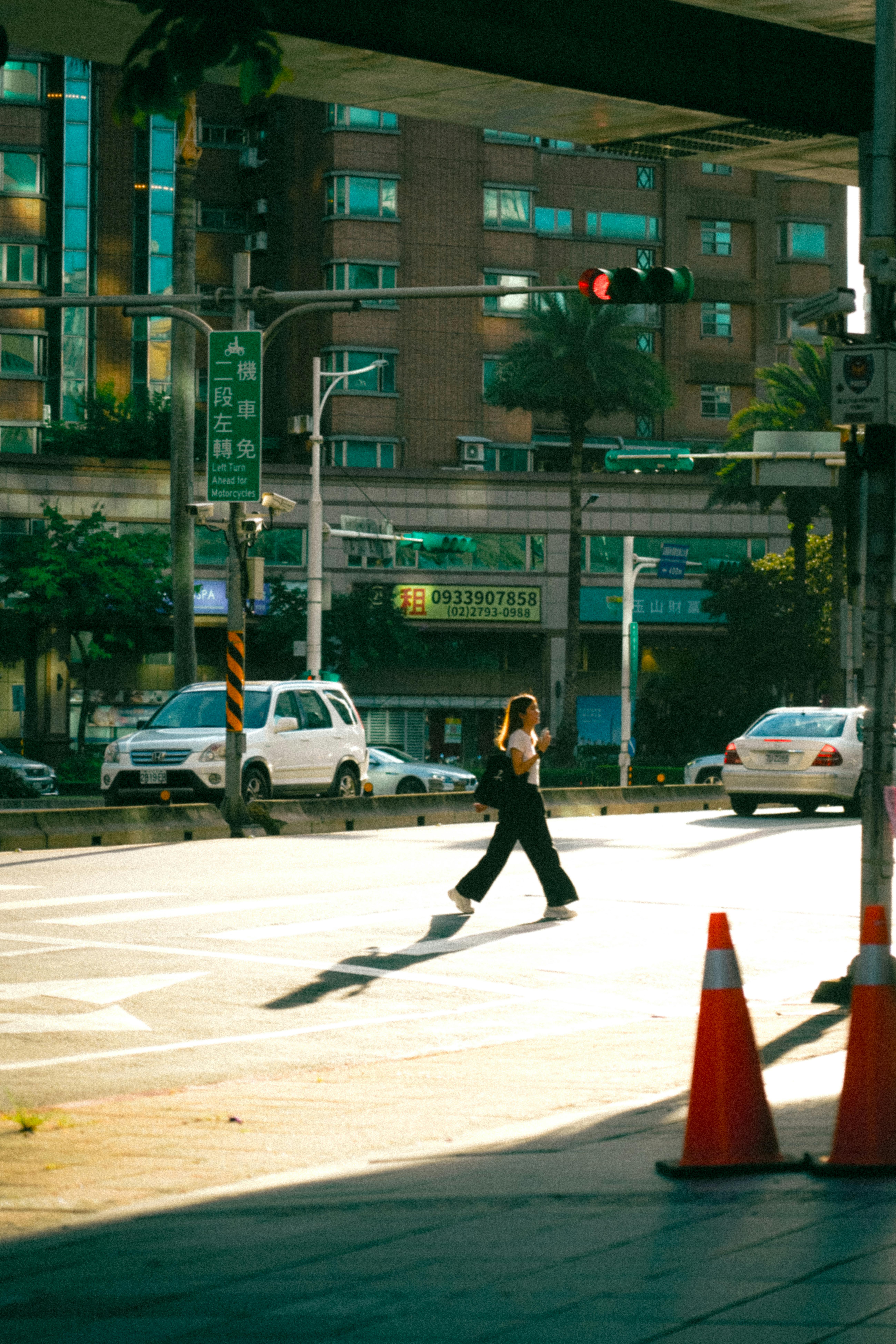 A woman walks across a sunlit city street with cars and buildings around.