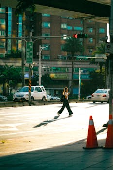A woman walks across a sunlit city street with cars and buildings around.