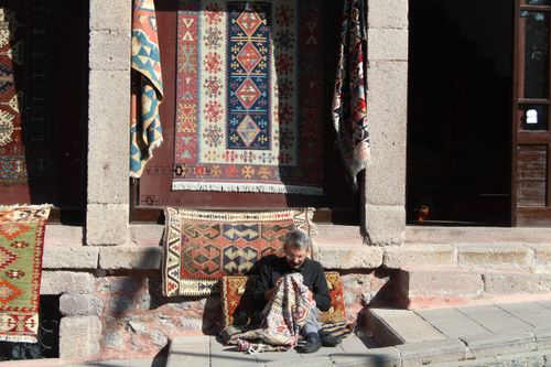 Free Man sitting with handmade carpets on a sunlit street, displaying traditional patterns and craftsmanship. Stock Photo