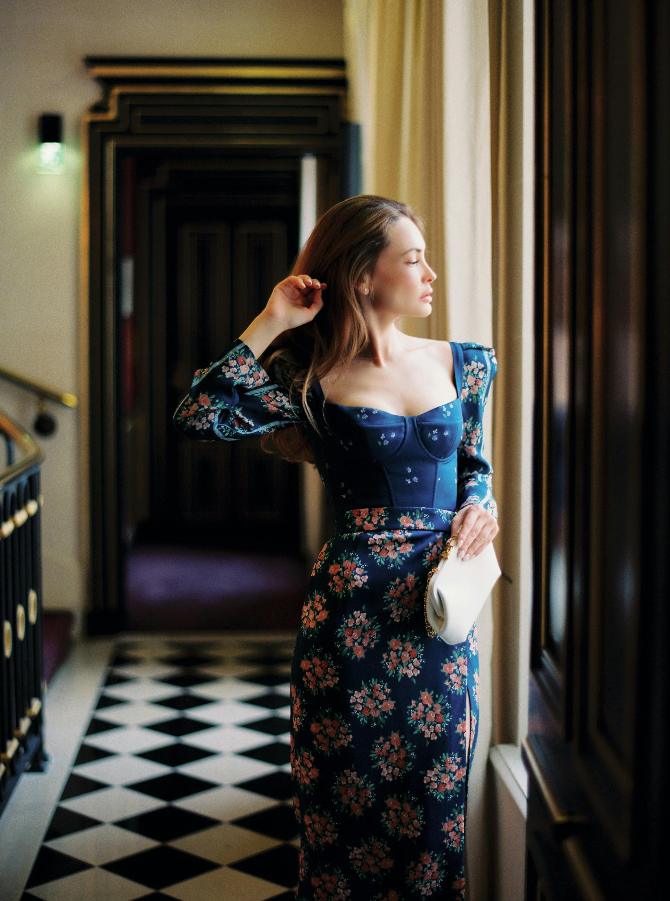 A stylish woman in a floral dress stands by a window, embodying elegance and sophistication.