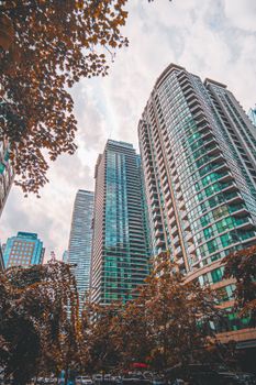 Low angle view of modern skyscrapers with autumn trees in an urban setting on a cloudy day.