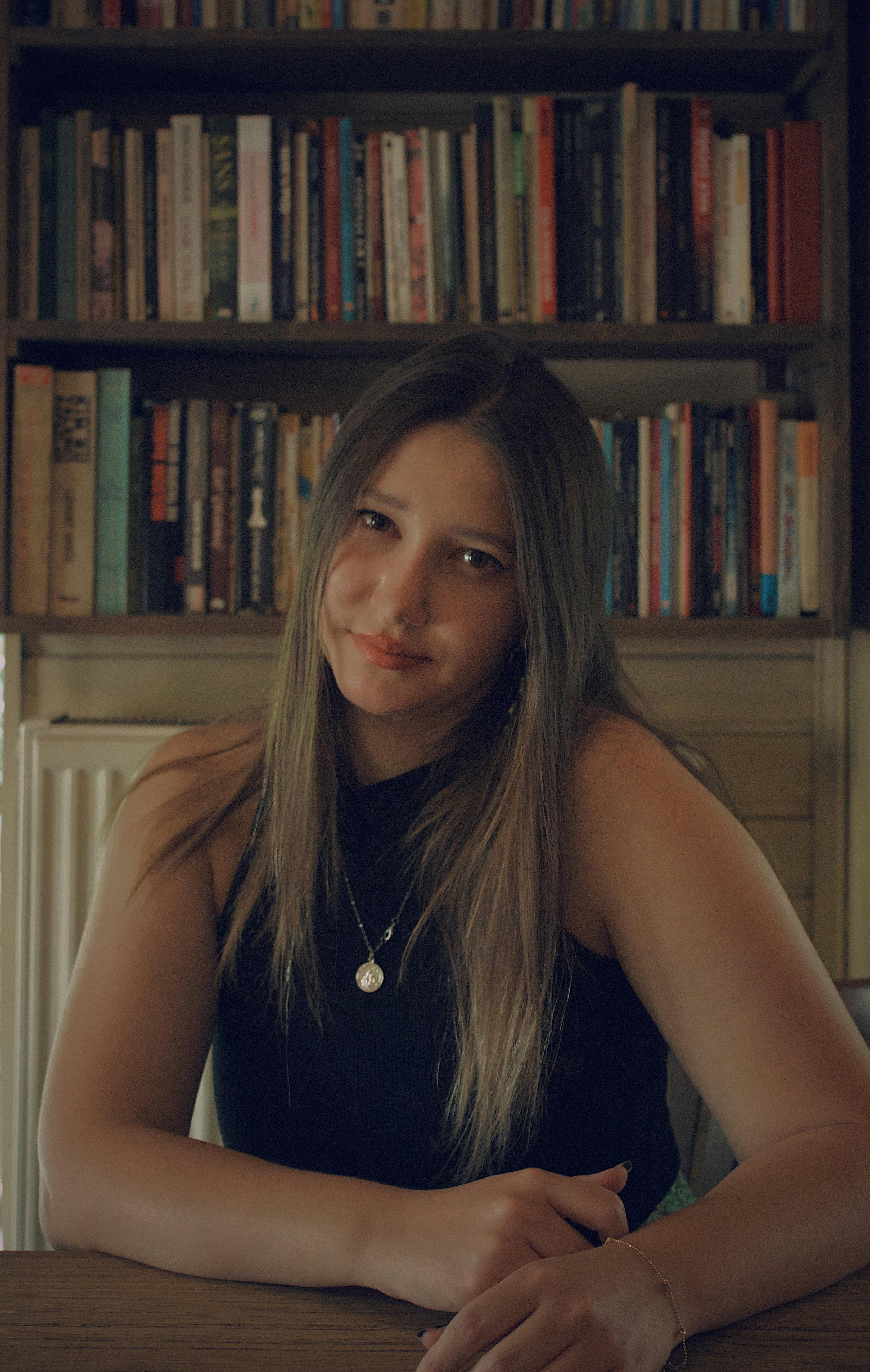 Portrait of a young woman sitting in a library with bookshelves in the background.