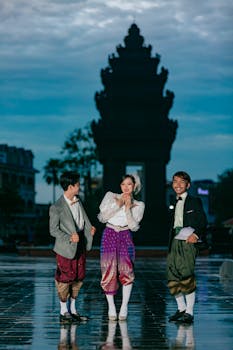 Three adults in traditional Cambodian clothing, smiling by a monument under a cloudy sky.