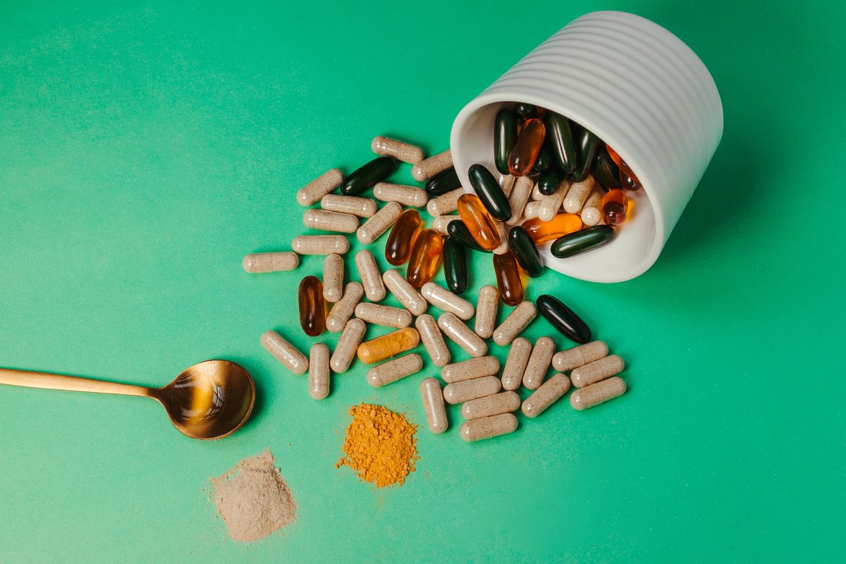 Capsules spilling from a white container beside small piles of loose supplement powder and a wooden spoon