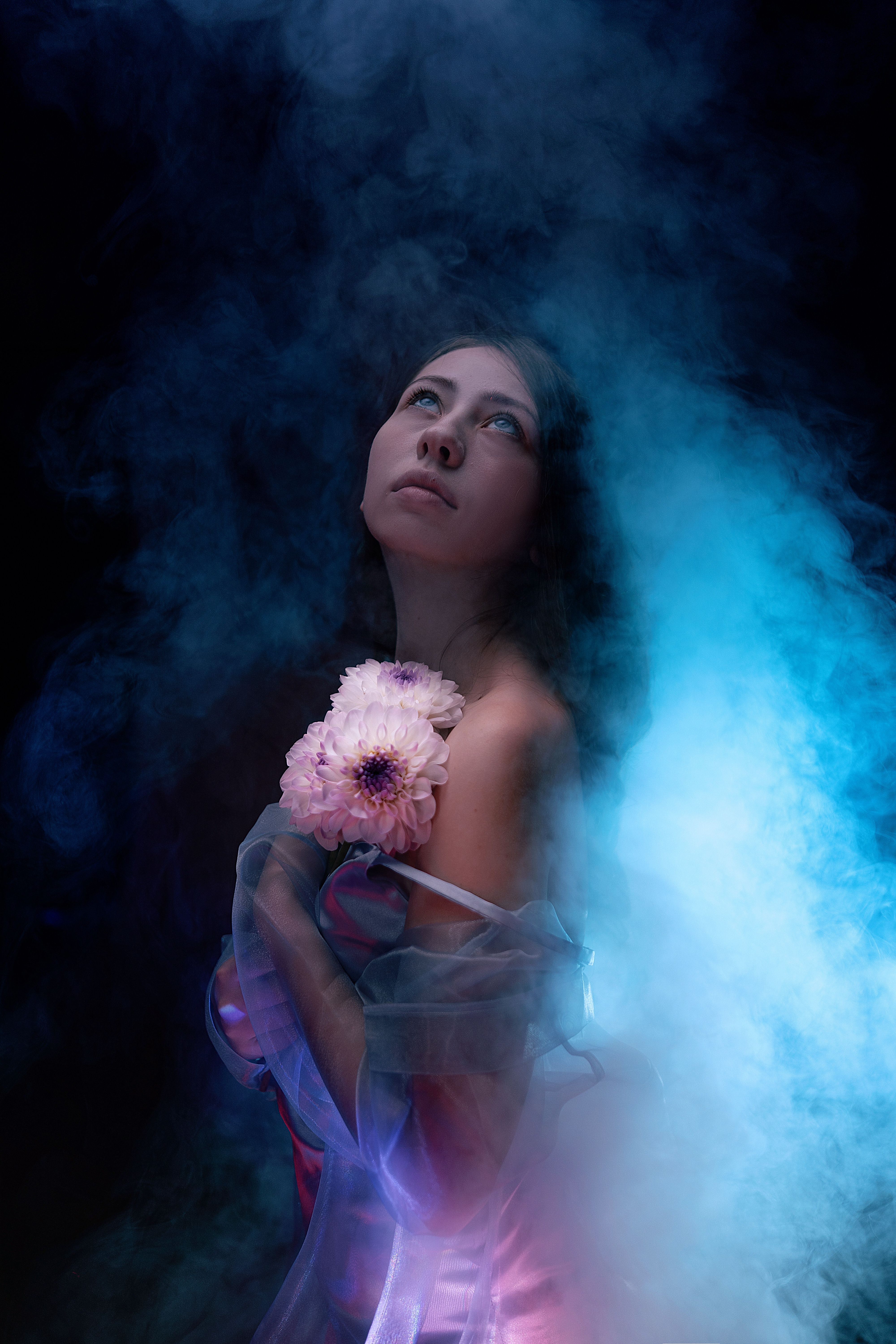 Dreamy portrait of a woman in soft blue and pink smoke holding a flower, with an ethereal and artistic aura.