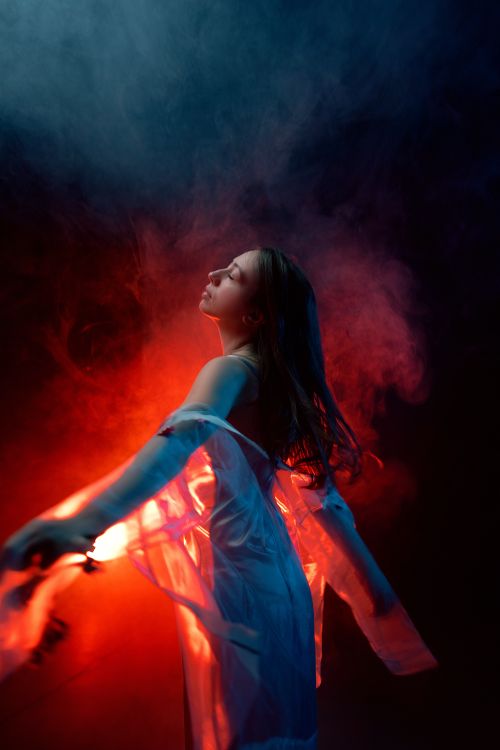 Free A young woman posing gracefully in a tulle dress enveloped in colorful fog and dramatic lighting. Stock Photo
