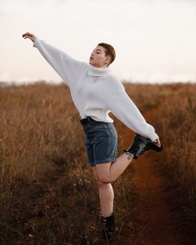 A young woman with short hair poses gracefully in a field wearing a white sweater and denim shorts.