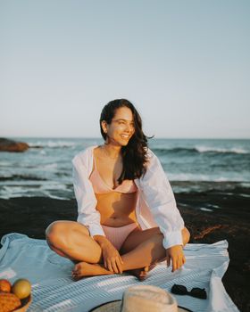 A woman in swimwear enjoys a sunny beach picnic by the sea, exuding relaxation and happiness.
