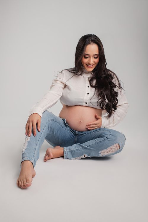 Free Happy pregnant woman with long hair sitting barefoot in casual clothes, gently touching her belly and smiling. Stock Photo