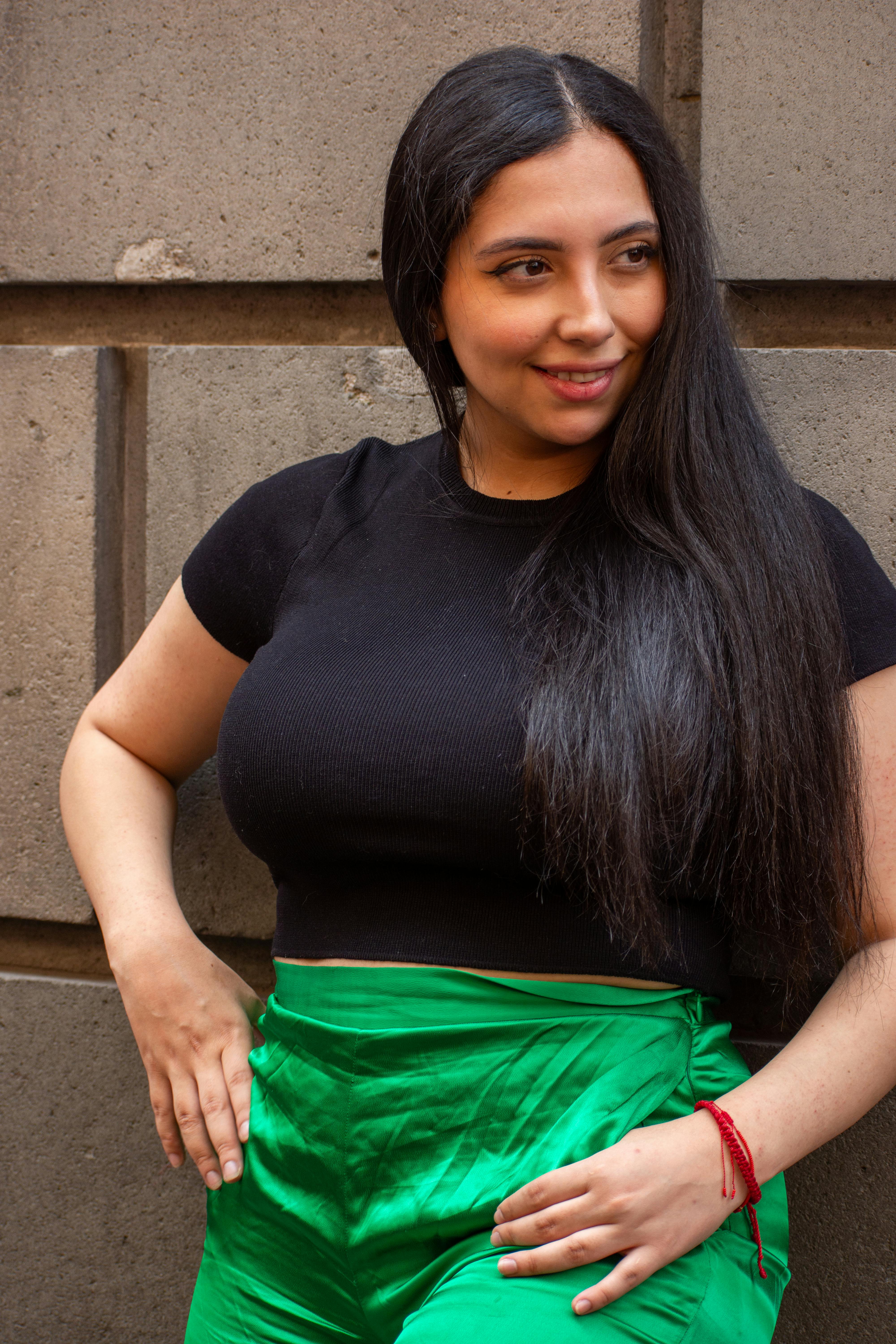 A young woman with long hair poses against a stone wall, wearing a black top and vibrant green pants.