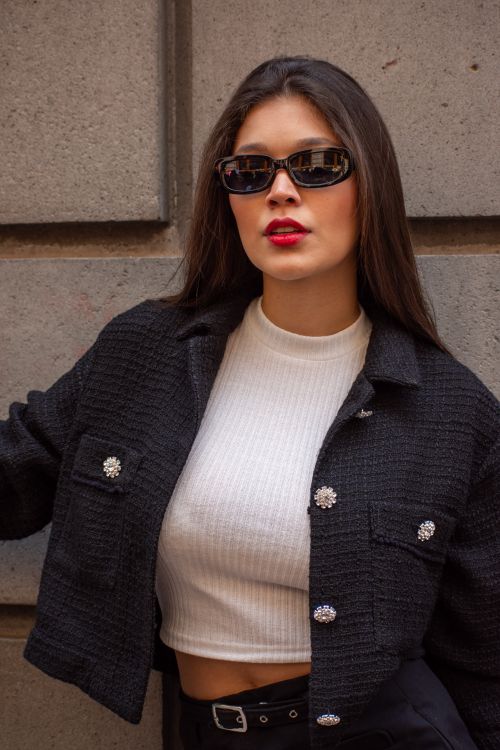 Free Stylish young woman wearing a black jacket and sunglasses posing confidently against a wall. Stock Photo