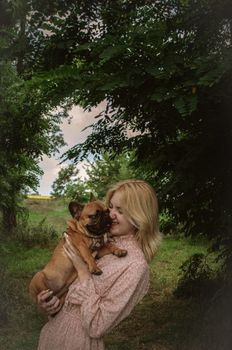 A joyful woman holds her French Bulldog in a lush green park setting.