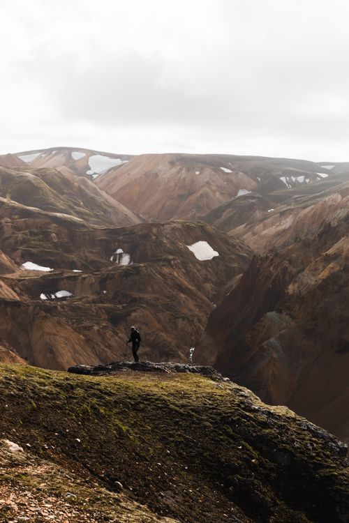 Free A lone hiker stands atop a hill, surrounded by dramatic mountainous terrain under a cloudy sky. Stock Photo