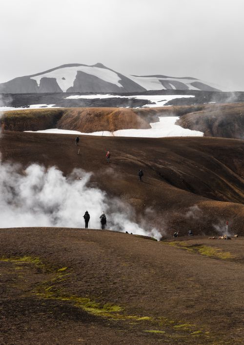 Free Hikers traverse steaming volcanic landscape with snow-capped mountains in the background. Stock Photo