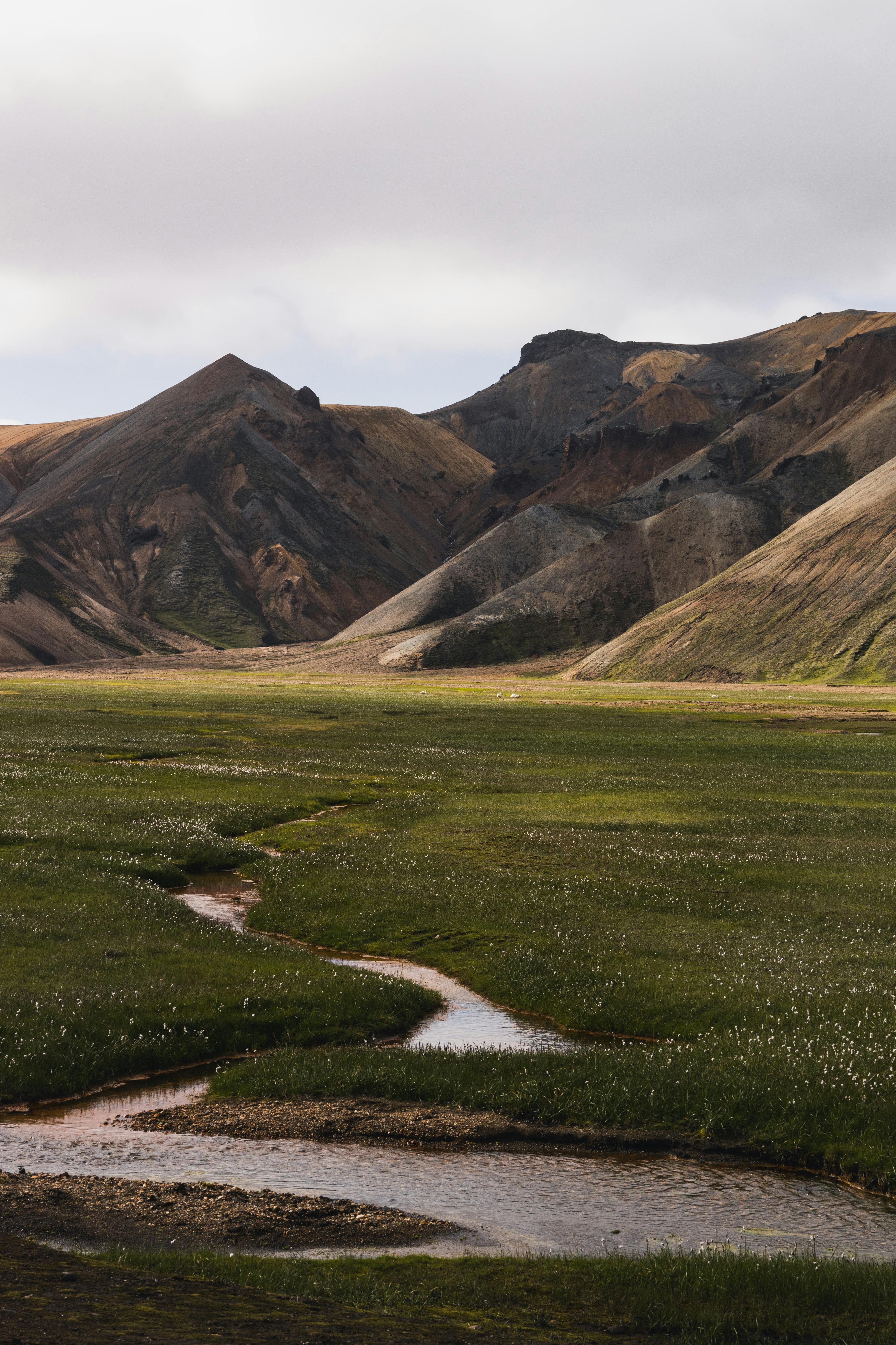 Explore a tranquil mountain valley with flowing stream and lush green grass under a cloudy sky.