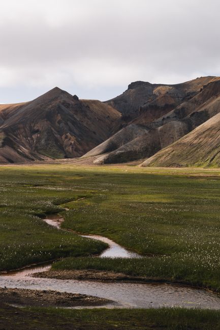 Explore a tranquil mountain valley with flowing stream and lush green grass under a cloudy sky.