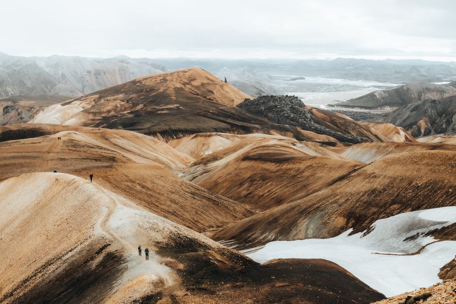 Expansive view of rugged, eroded mountains with hiking trail, snowy patches, and distant valley.