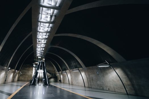 Wide-angle view of a futuristic and empty underground metro station with escalators and arched ceilings.