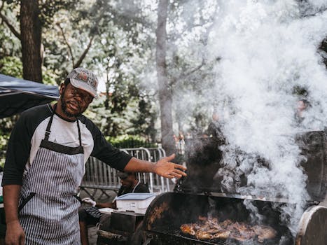 A man grilling meat outdoors with smoke rising from the barbecue.