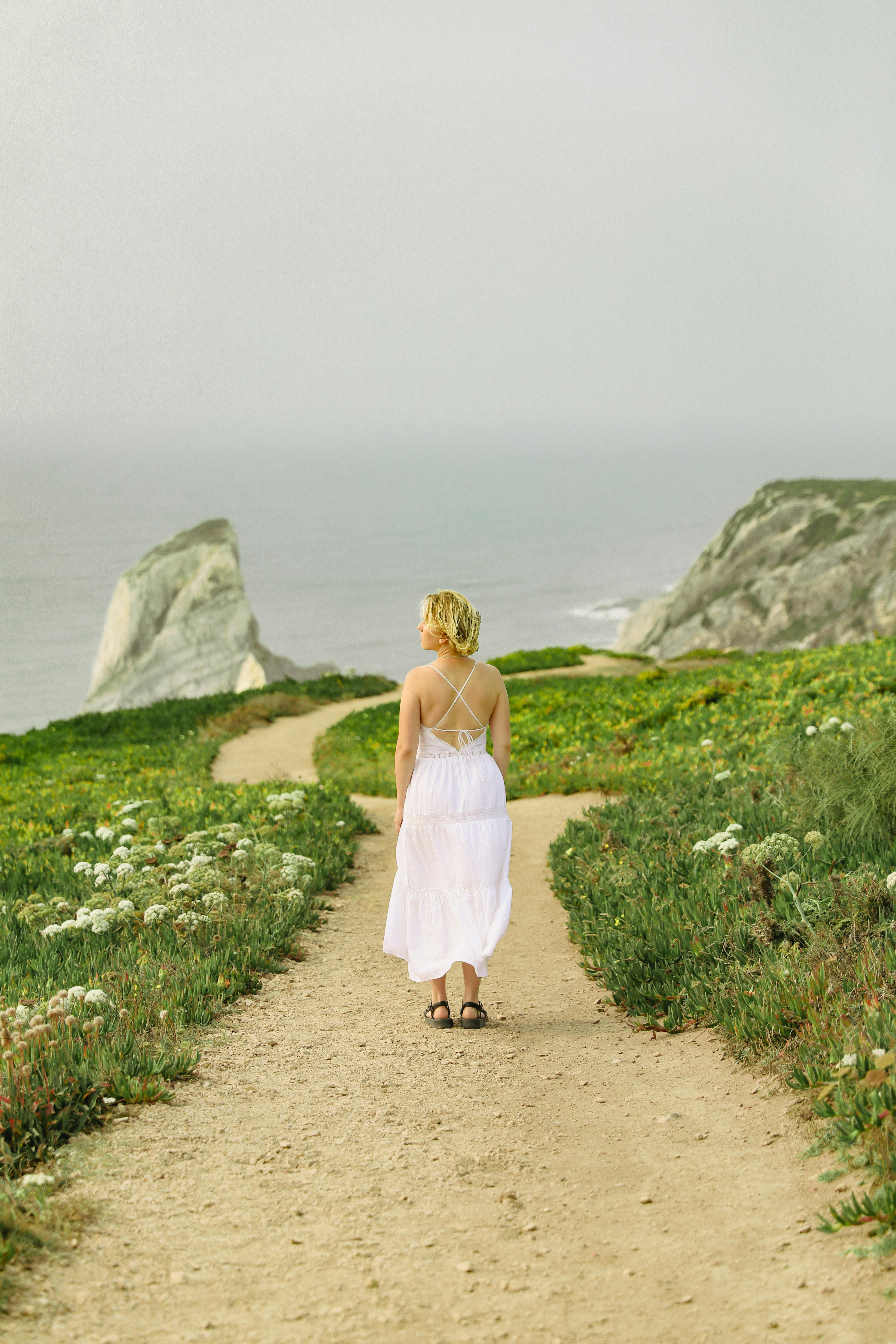 A woman walks on a path overlooking rocky cliffs and the ocean, embodying tranquility and summer vibes.