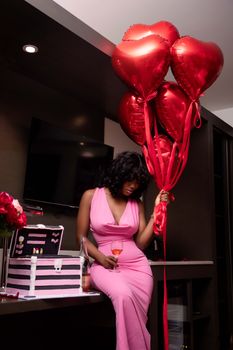 Elegant woman in a pink dress celebrating with red heart balloons indoors.