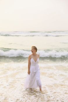 Elegant woman walking on the shore in a flowing white dress, enjoying the waves.