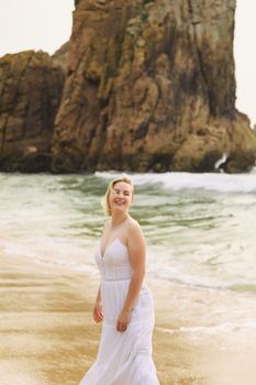 Blonde woman in a white dress posing joyfully on a sandy beach with rock formations.