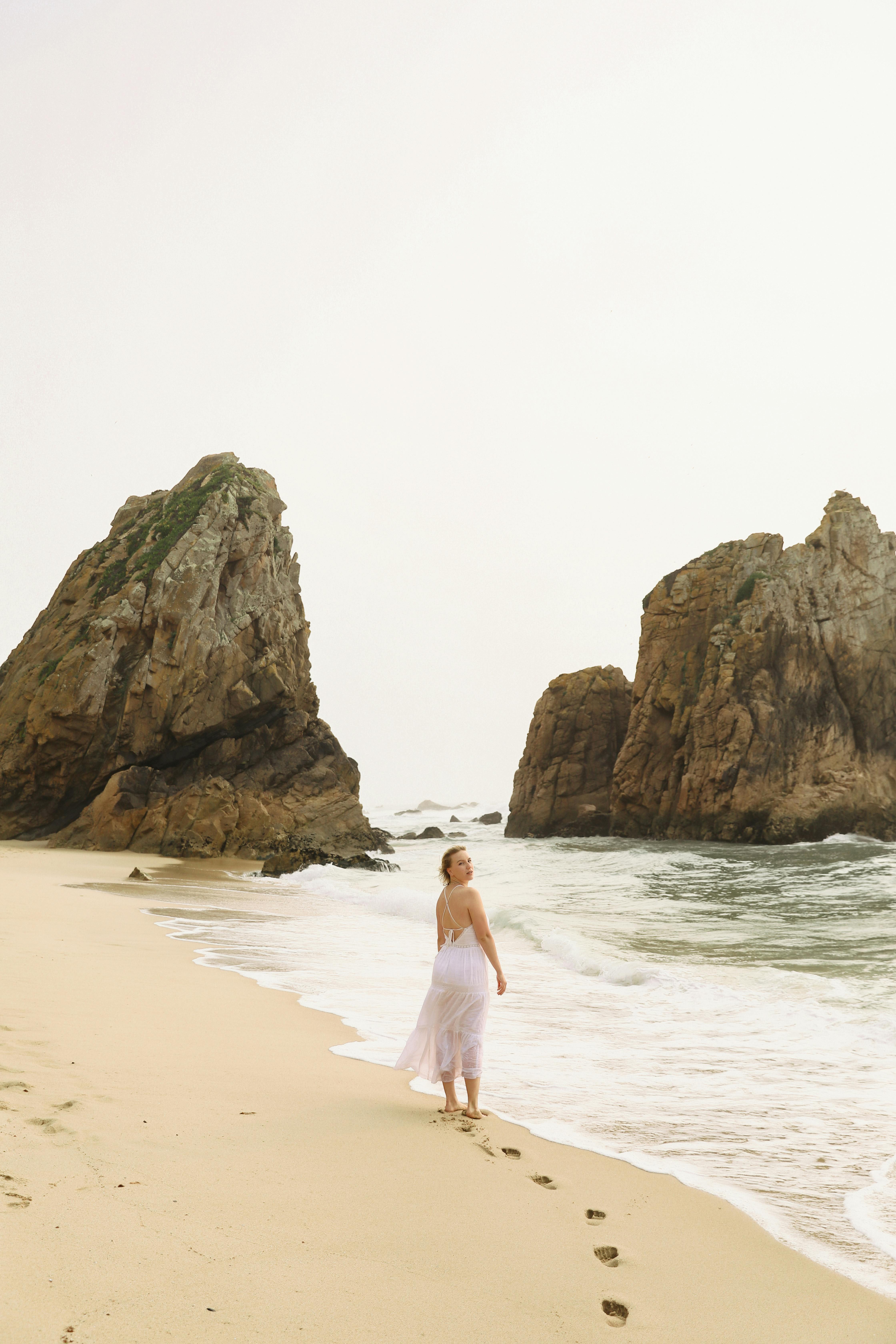 Woman in white dress walking along scenic Ursa Beach, Portugal, with majestic rock formations.
