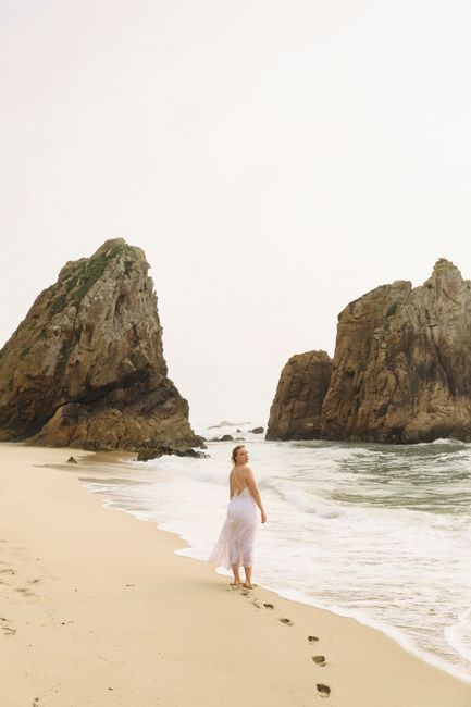 Woman in white dress walking along scenic Ursa Beach, Portugal, with majestic rock formations.