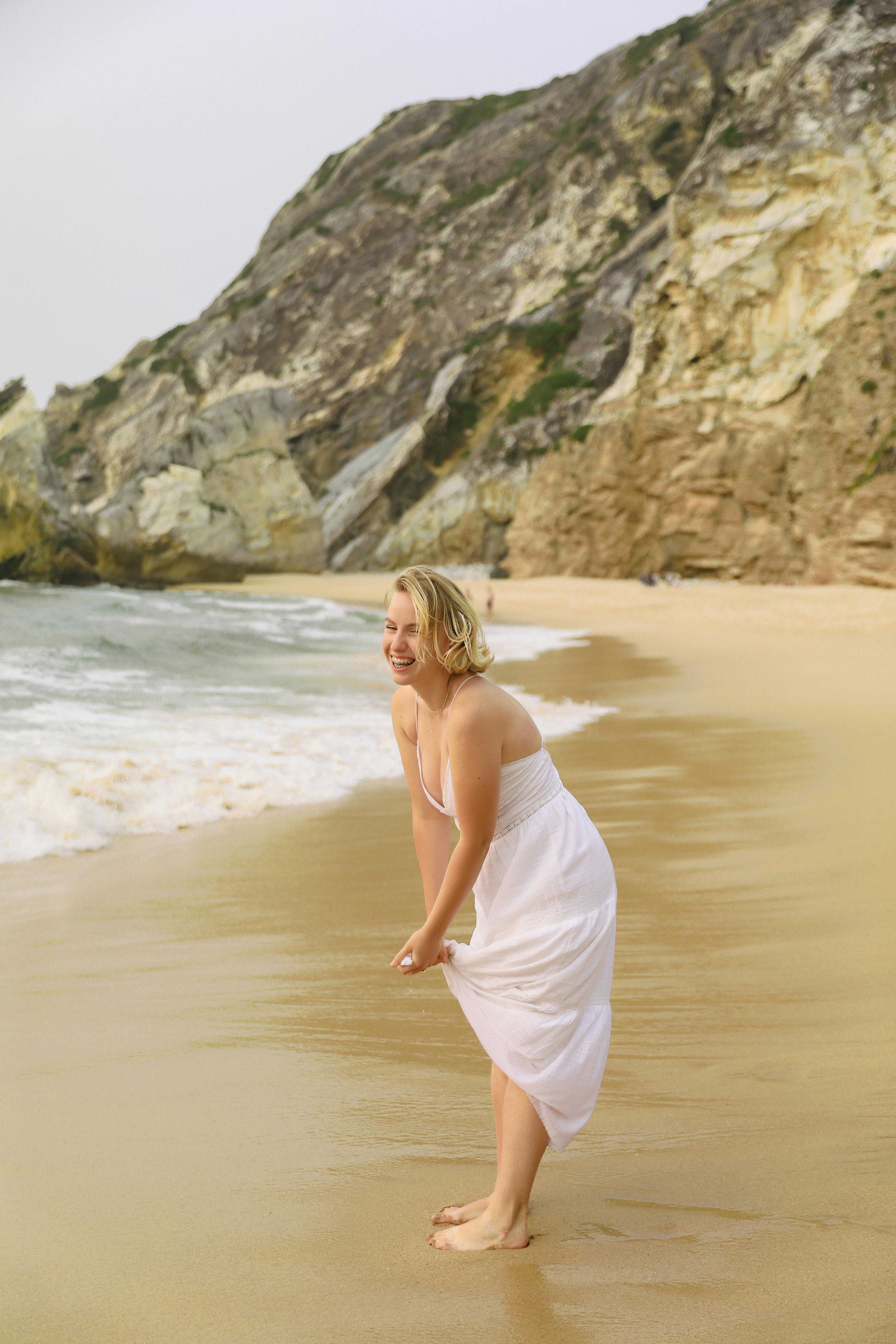 Smiling blonde woman in white dress enjoying a day at a sandy beach with rocky cliffs.