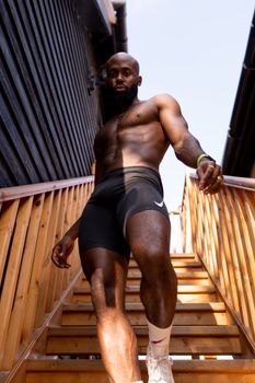 A shirtless, muscular man in sportswear ascending wooden stairs outdoors, highlighting fitness.