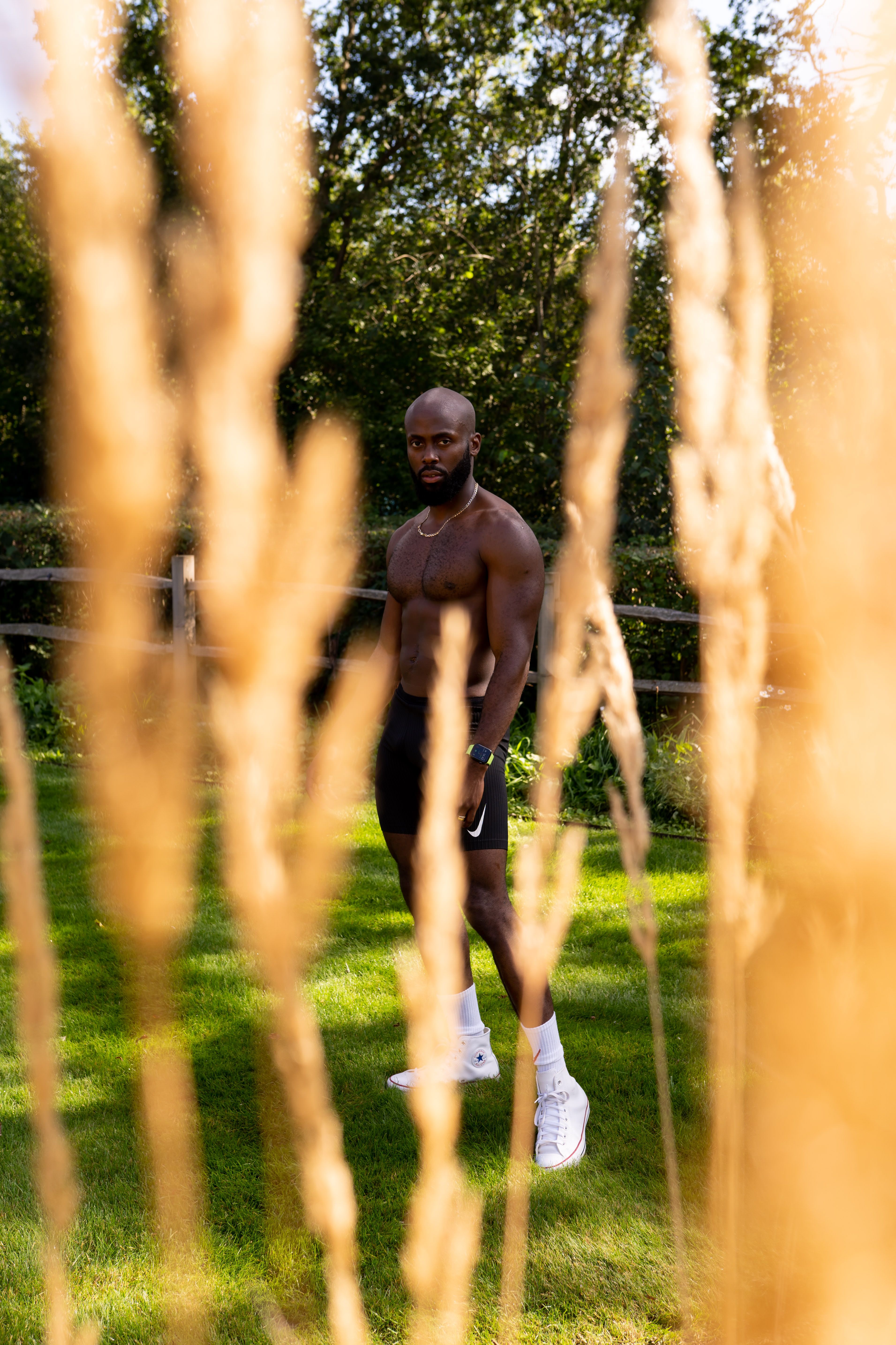A shirtless athletic man stands outdoors in a grassy area framed by tall plants.