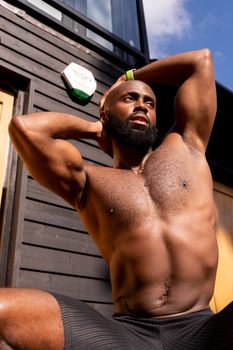 Muscular man posing shirtless outdoors against a wooden wall, exhibiting strength and confidence.