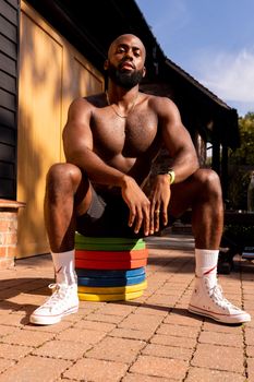 Shirtless bodybuilder sitting on colorful weight plates outdoors in a sunlit setting.
