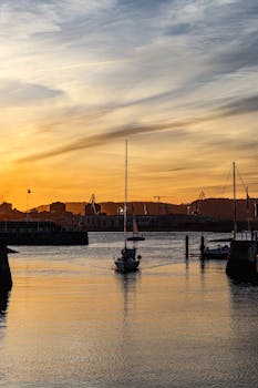 A serene sailboat glides through Gijón harbor during a vibrant sunset, capturing a peaceful maritime scene.