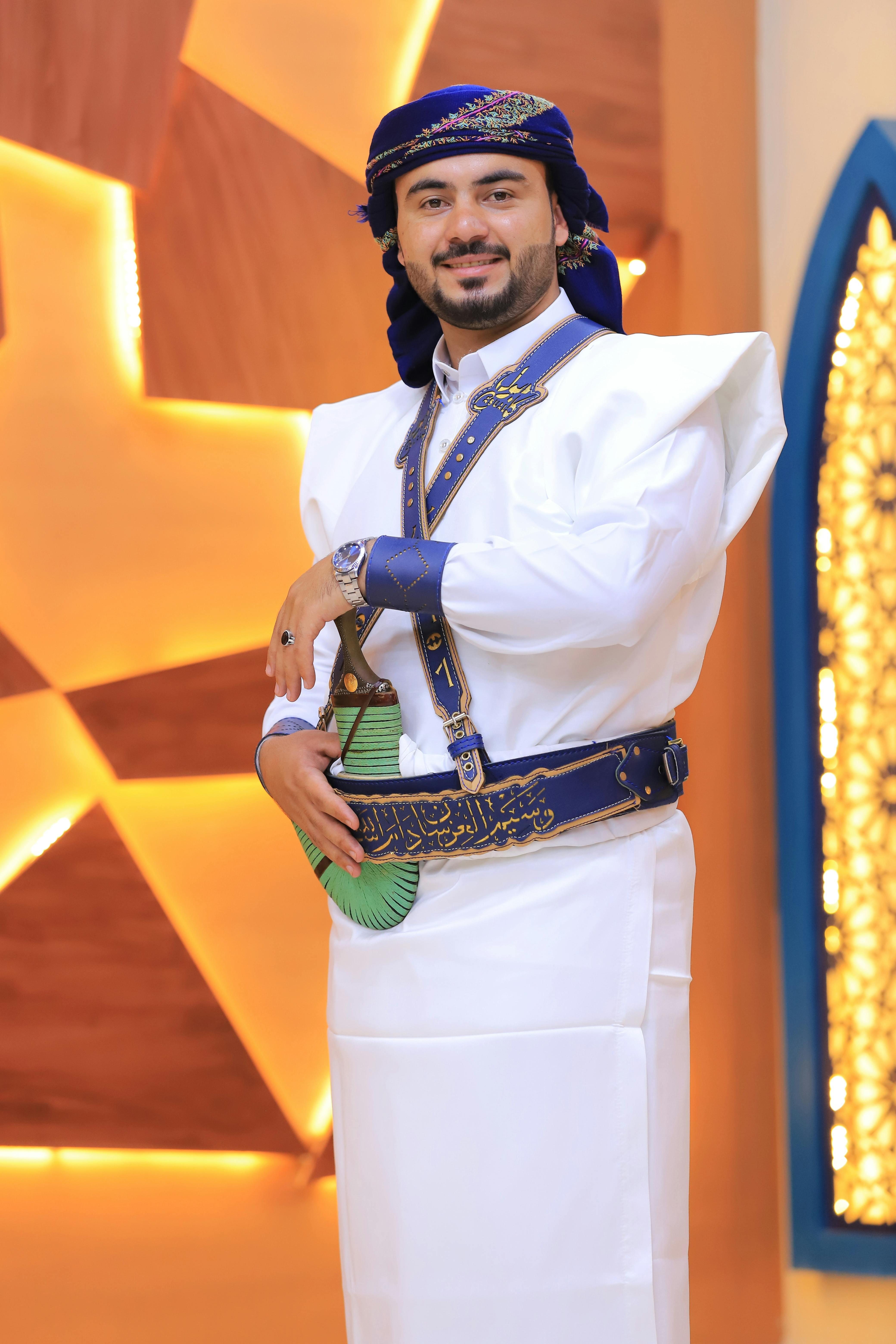 Young man in traditional clothing smiles indoors with decorative background.